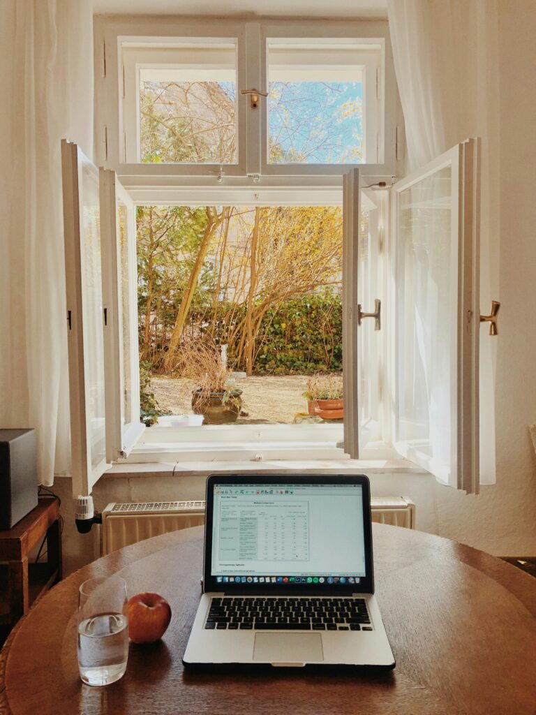 Cozy home office setup with laptop on desk facing an open window offering a view of a garden.